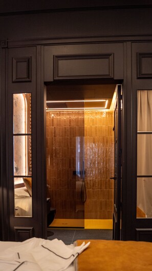 A hotel room with a wooden floor, a bed with white bedding, a bathroom with a shower and toilet, and a large window with curtains. - Boada Riera (Terrassa)