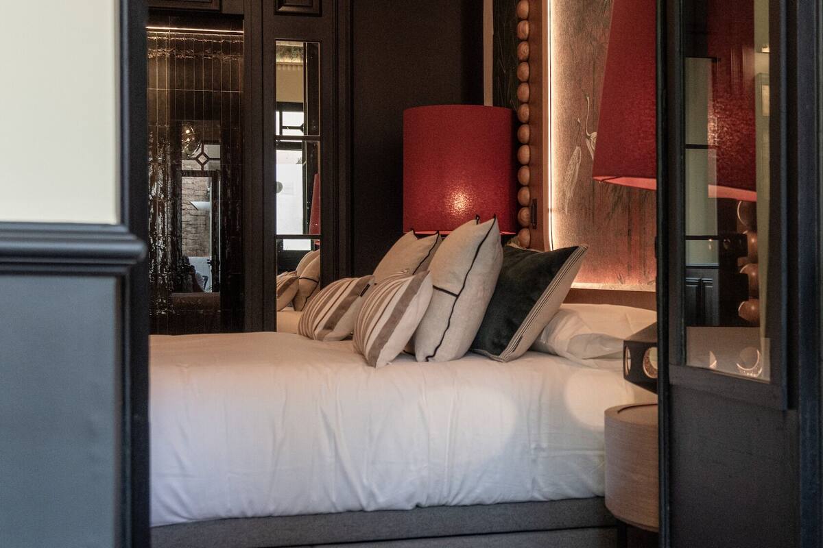 Una cama bien hecha con varios almohadones, un objeto cilíndrico rojo y un espejo que refleja parte de la habitación.