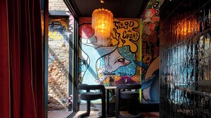 A dining area with a mural, two black chairs, a table, and a chandelier. - Boada Riera (Terrassa)