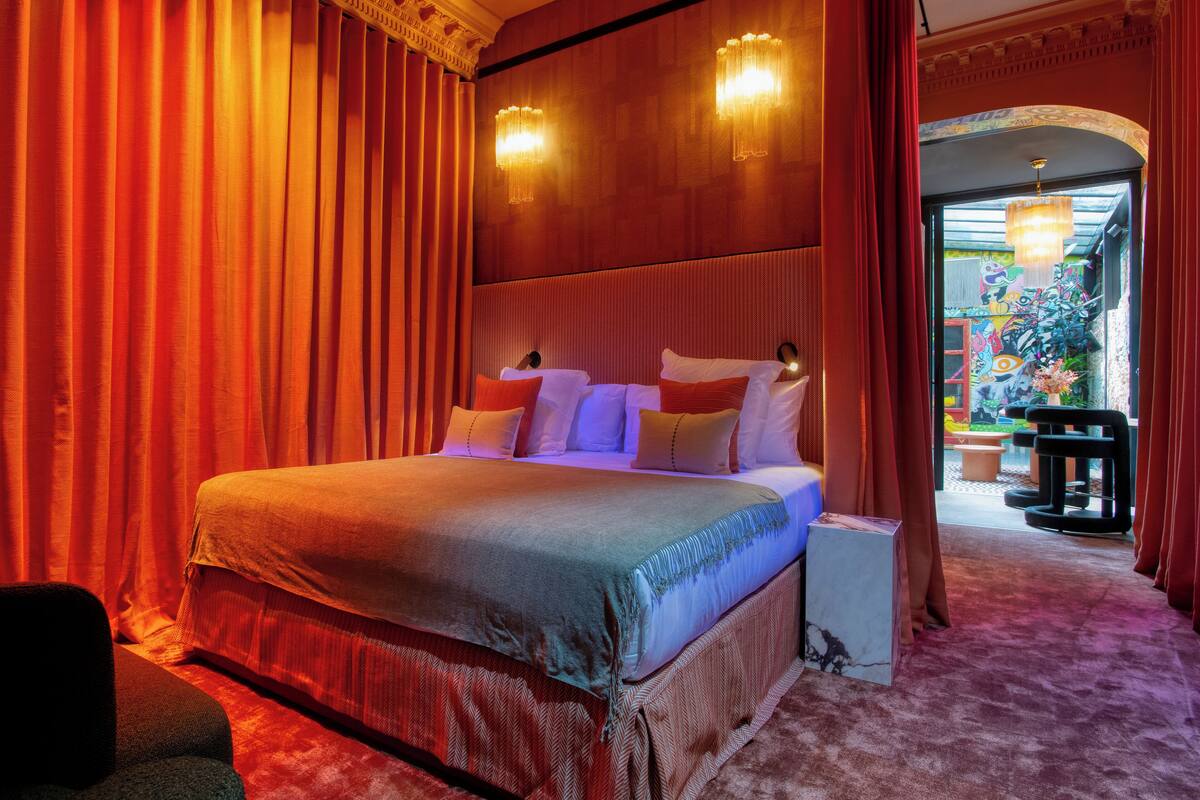 Un dormitorio con una cama grande, cortinas rojas y vistas a un espacio exterior con plantas y un mural colorido.