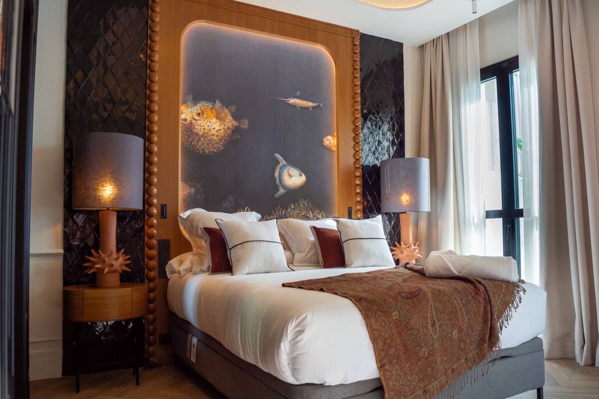 Una habitación de hotel moderna con una cama grande, dos mesitas de noche con lámparas, un mural decorativo y un ventanal con cortinas.