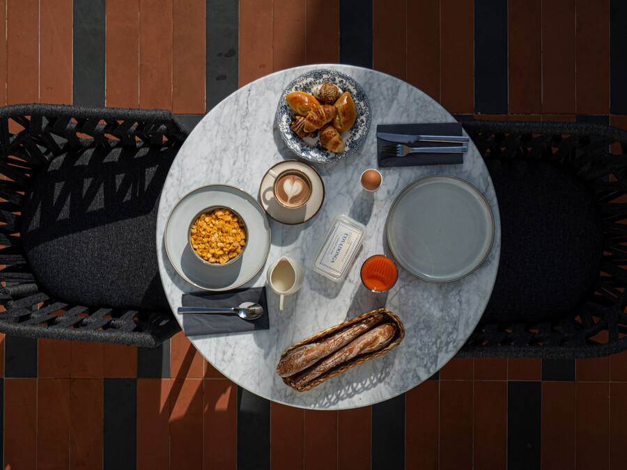 A round table with a white marble top, surrounded by black woven chairs. On the table, there is a plate with croissants, a bowl of cereal, a cup of coffee, a glass of orange juice, and a baguette.
