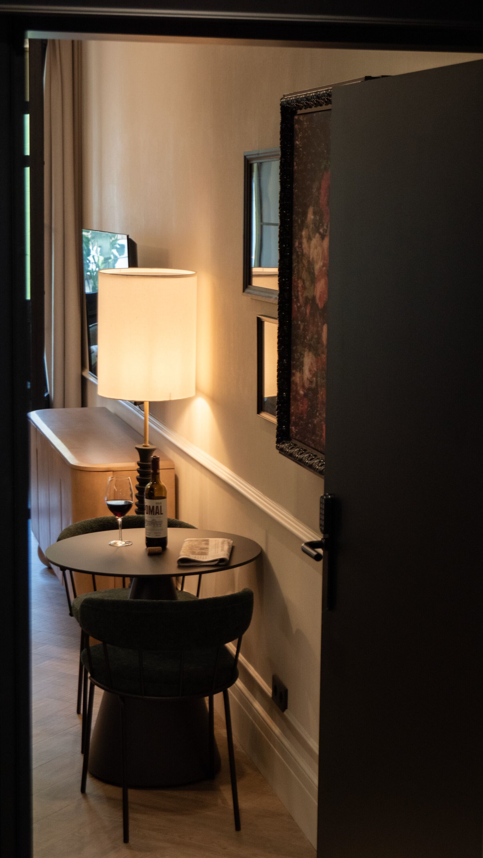 A small dining area with a table, chairs, a lamp, a bottle of wine, and a glass of wine.