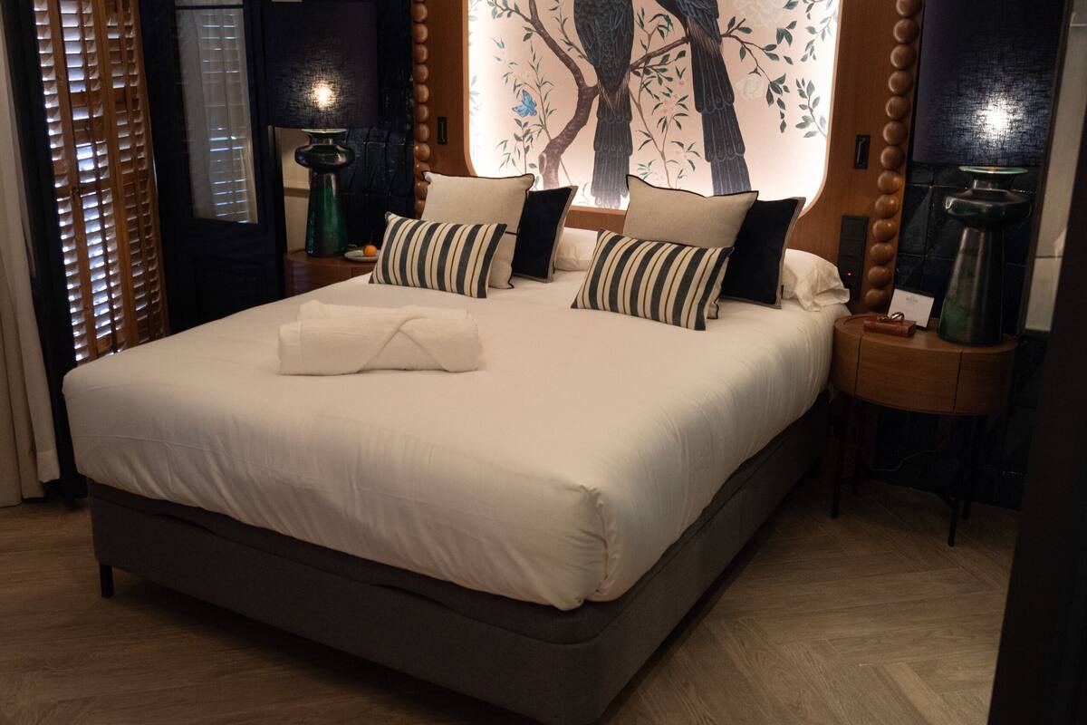 Una cama grande con edredón blanco, almohadas a rayas y una manta a juego. Hay dos mesitas de noche con lámparas y una obra de arte en la pared que representa un árbol.