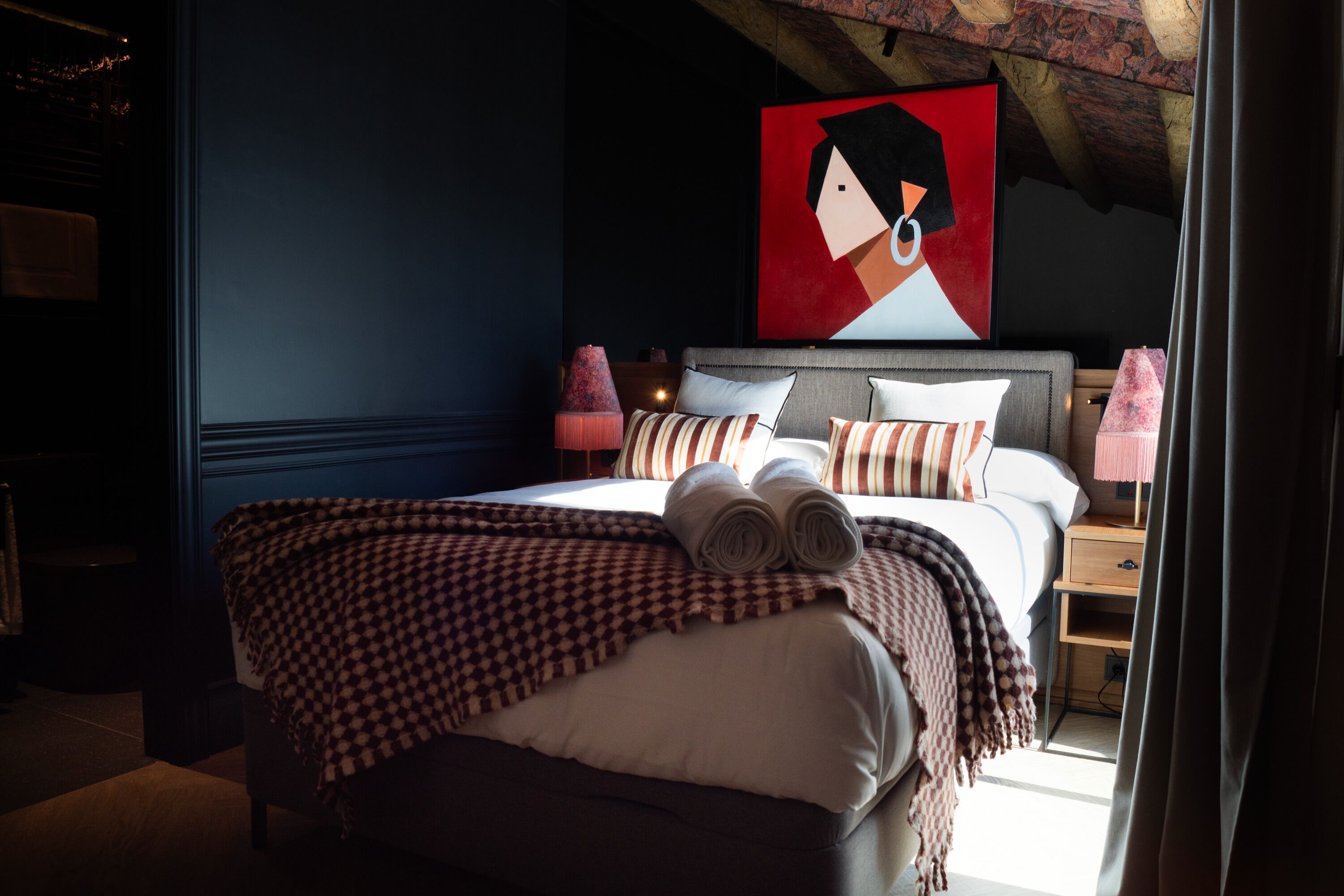 Un lit bien fait, avec une couverture à carreaux, un oreiller et des serviettes enroulées. Un tableau est accroché au mur au-dessus du lit, et une table de chevet avec une lampe se trouve à côté.