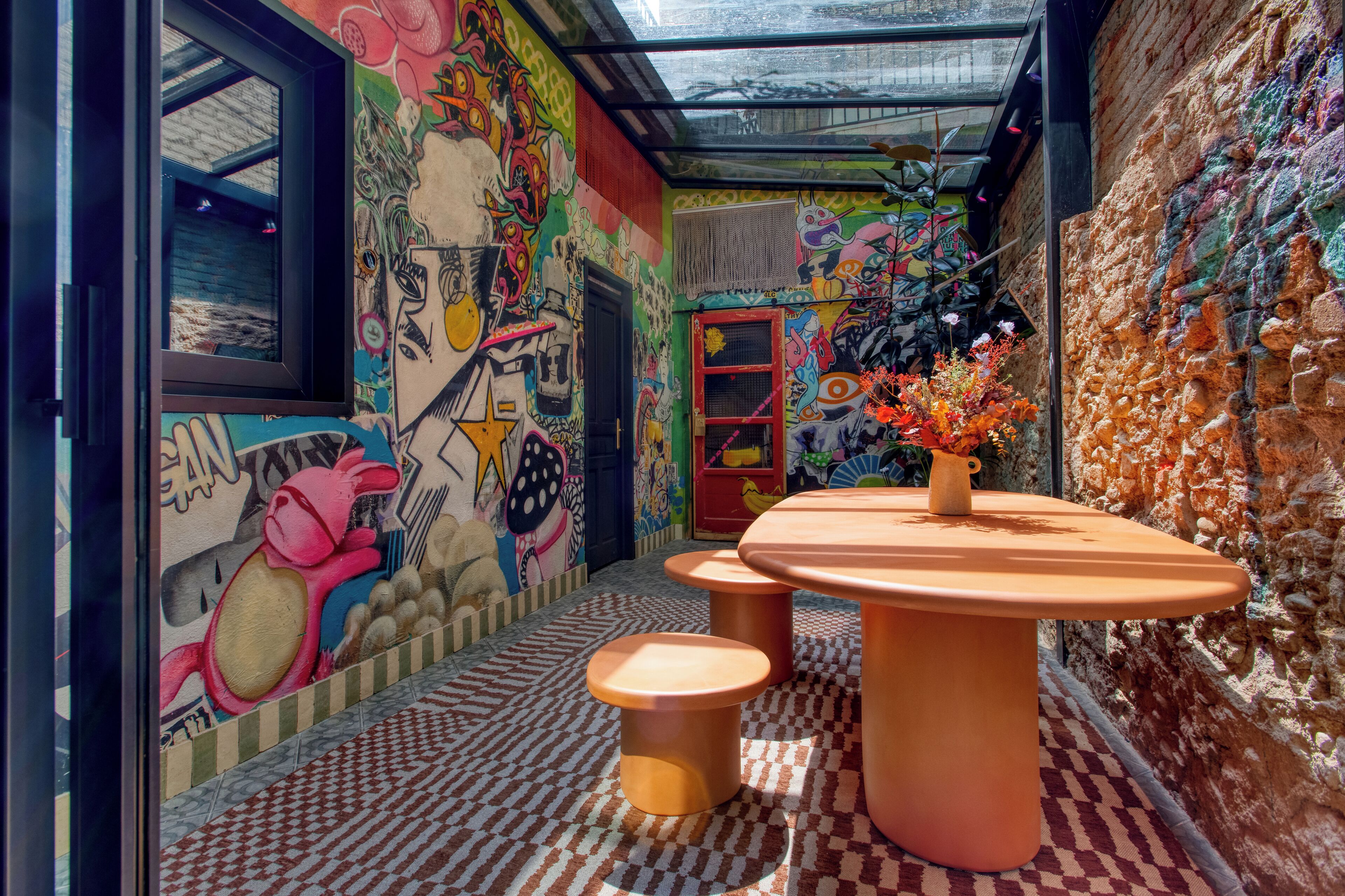 Un pasillo estrecho con grafitis coloridos en las paredes, suelo a cuadros y una mesa con taburetes.