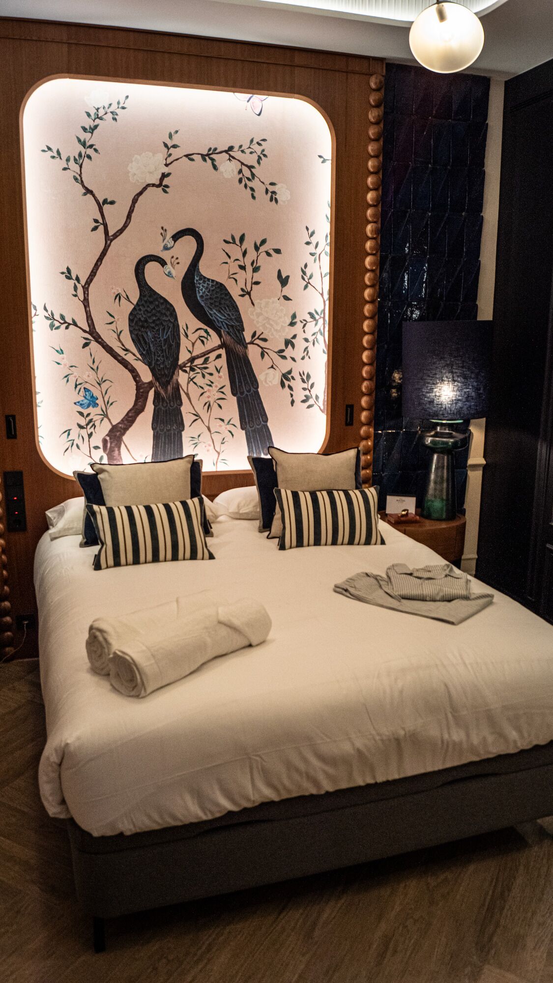 A neatly made bed with pillows and a rolled towel, a wall with a floral and bird mural, a lamp, and a dark-colored nightstand.