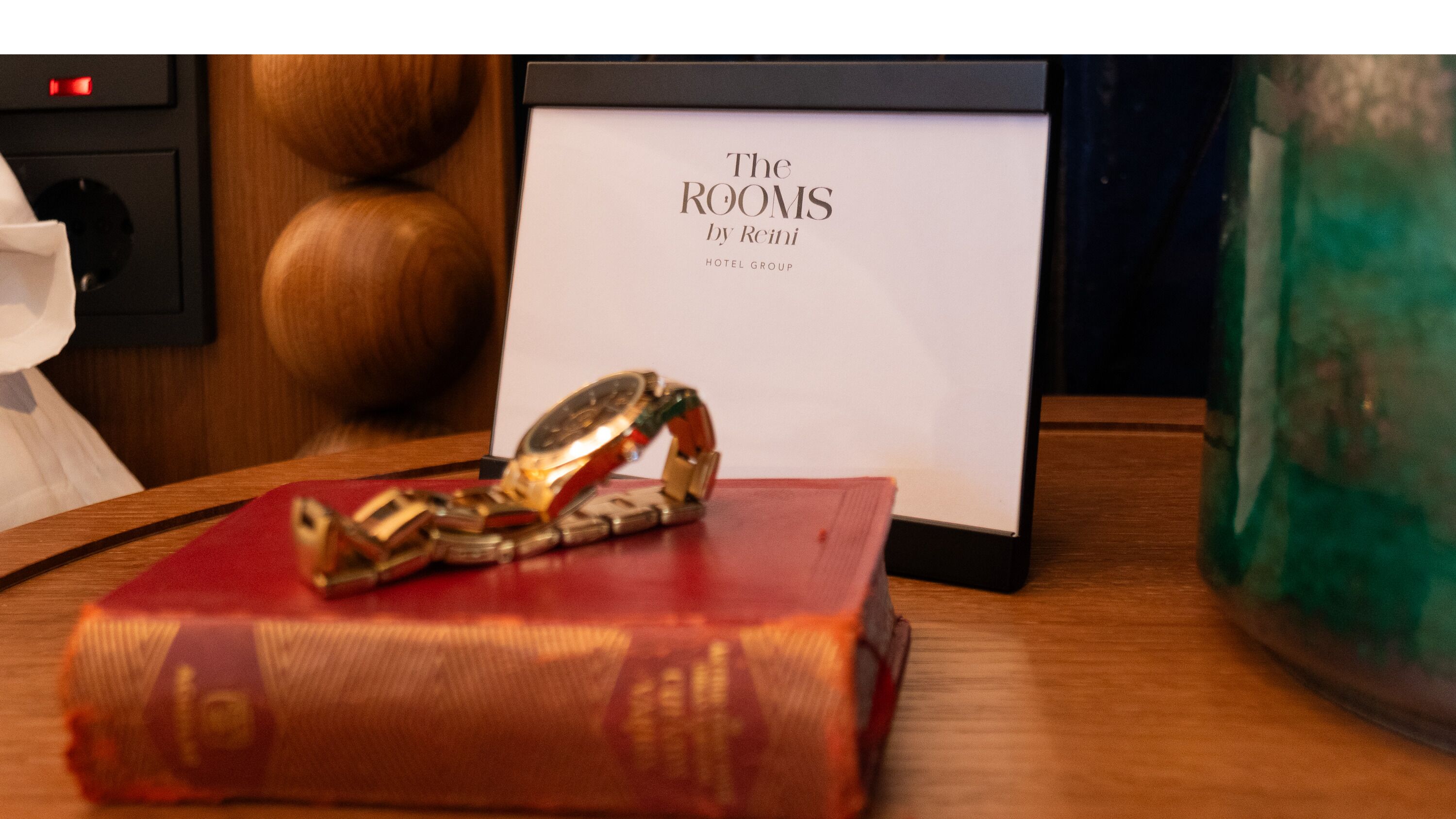 A book titled "The Rooms by Reini" on a wooden table with a watch and a framed sign.