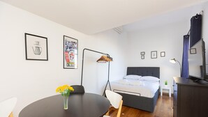 Een moderne slaapkamer met een bed, nachtkastje en een televisie. Er staat een klein tafeltje met een bloevas en een stoel. De kamer heeft een houten vloer en witte wanden.