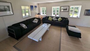 A modern living room with a sectional sofa, a white bench, and a coffee table. There are three windows providing natural light. - 5 Star Holiday Home in Vesterborg (Vesterborg)