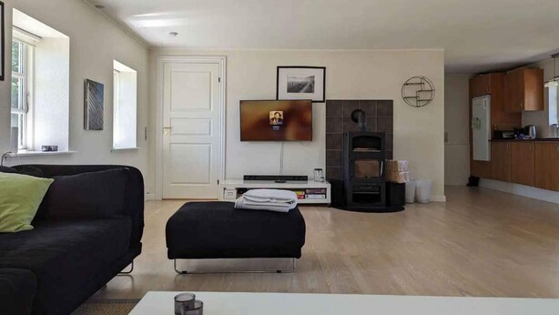 A modern living room with a black sofa, a white coffee table, a fireplace, and a kitchen area with wooden cabinets. - 5 Star Holiday Home in Vesterborg (Vesterborg)