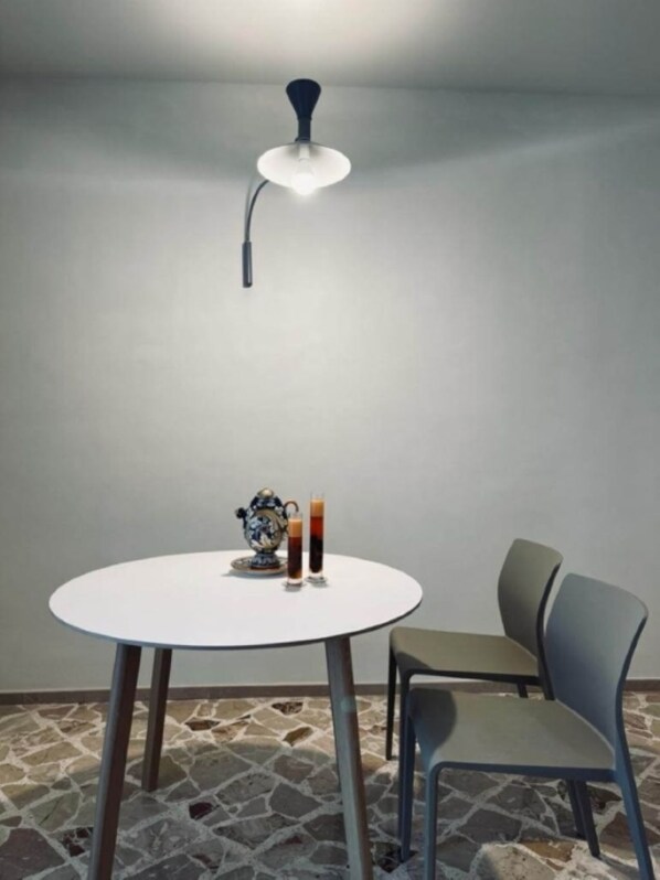 Une table ronde avec un plateau blanc et des pieds en métal, accompagnée d’une chaise avec un siège et un dossier gris. Une théière décorative et deux verres contenant un liquide foncé, probablement du café ou du thé, sont posés sur la table. Un luminaire suspendu de style moderne est accroché au-dessus de la table. Le sol est recouvert de carreaux de pierre à motifs.