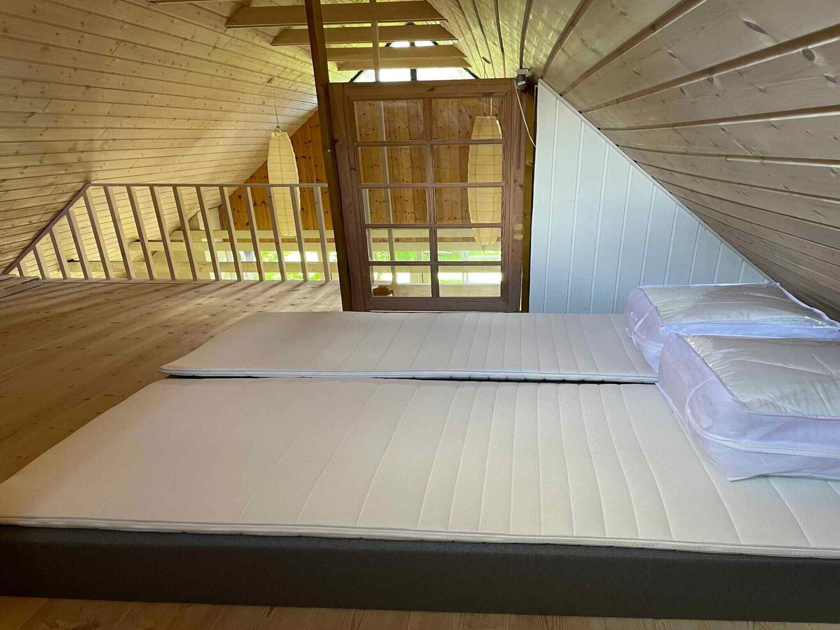 A loft bedroom with a wooden sloped ceiling, a mattress, and a view through a wooden door.