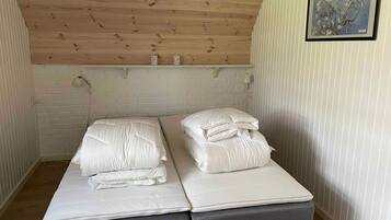 Um quarto pequeno e acolhedor, com teto de madeira e parede de tijolos brancos. Há uma cama com roupa de cama branca e uma mesa pequena com um abajur.