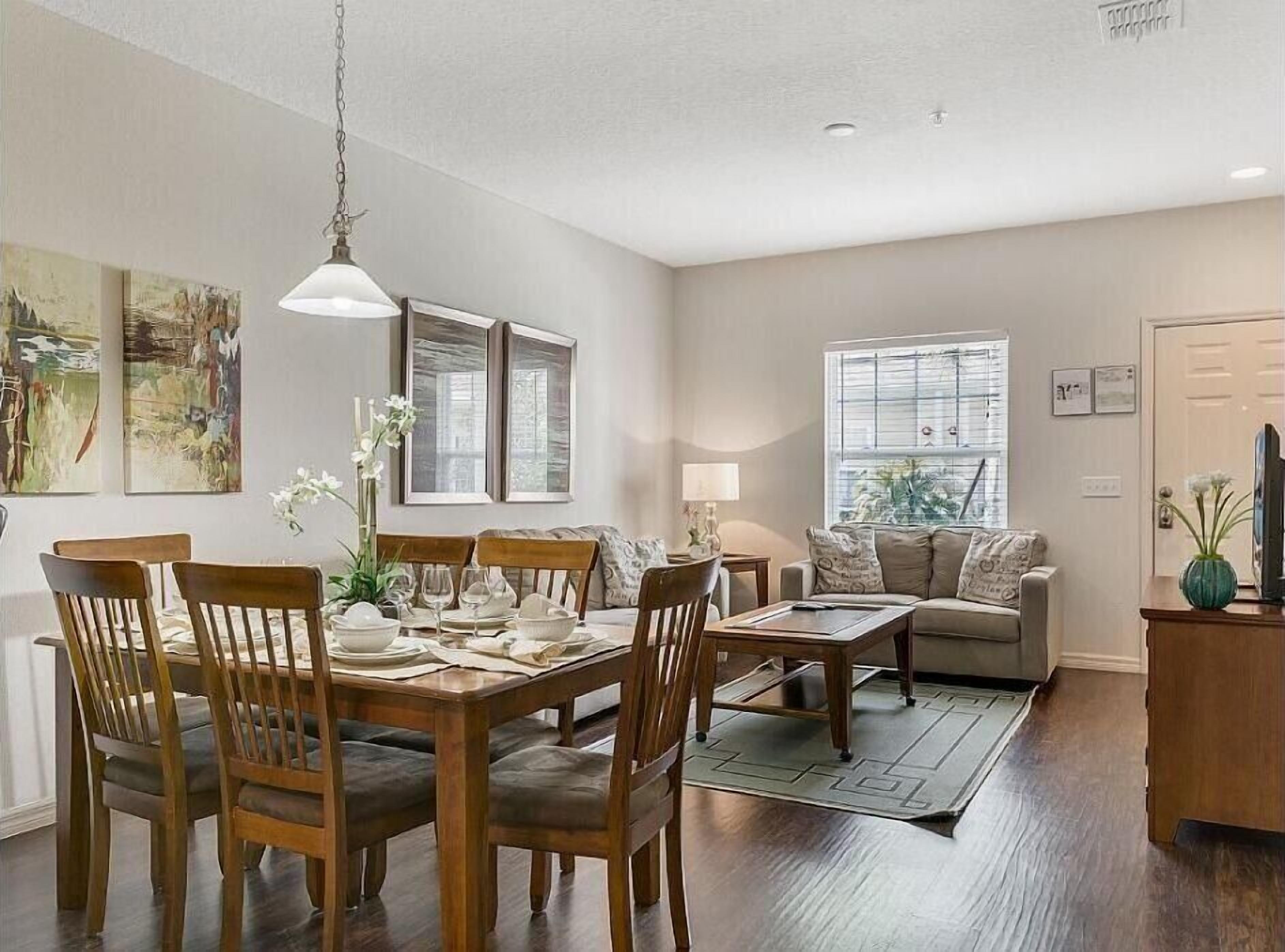 A dining area with a wooden table and chairs, a hanging light fixture, and a window with blinds.