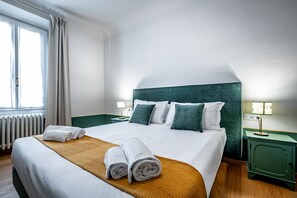 Ein ordentlich bezogenes Bett mit weißen Leinen, einem Holzkopfstück und grünen Kissen. Es gibt zwei Nachttische mit Lampen und ein Fenster mit Vorhängen.