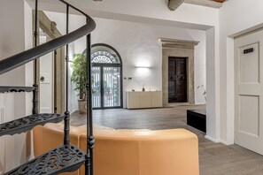Una escalera con barandales ornamentados, un banco con cojín color beige, piso de madera, una puerta blanca y un amplio ventanal de arco con reja de metal.