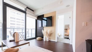 Quarto de hotel moderno com uma grande janela, televisão de tela plana na parede, piso de madeira e vista para o horizonte urbano.