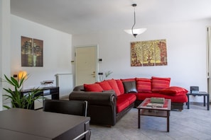 A living room with a sofa, a coffee table, a dining table, and chairs. There is a radiator, a door, and artwork on the walls.