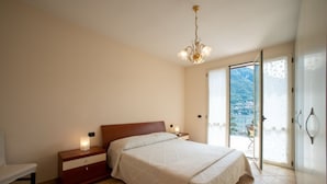 Une chambre à coucher avec un lit, des tables de chevet, une commode, un lustre et une vue sur l’extérieur, visible à travers une porte vitrée.