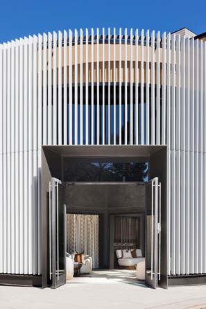 Een modern gebouw met een witte gevel, grote glazen deuren en een zichtbaar interieur met een bank en gordijnen.