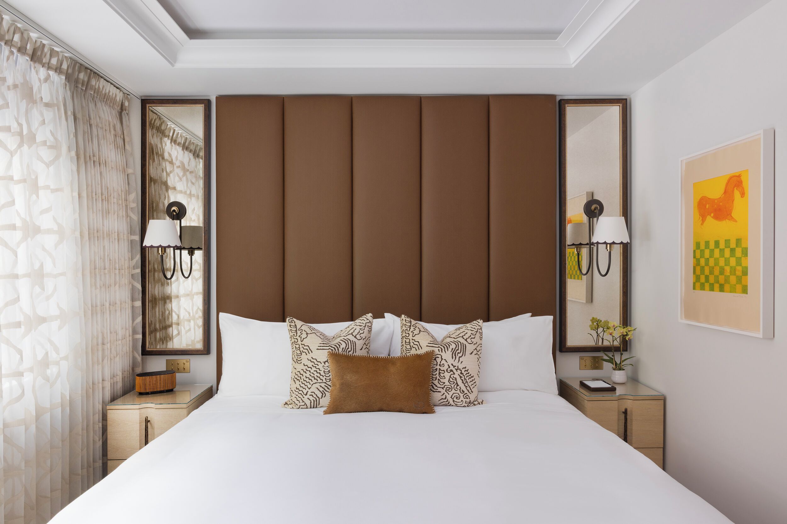 A hotel room with a large bed, two bedside tables, a headboard, a mirror, and a wall-mounted artwork.