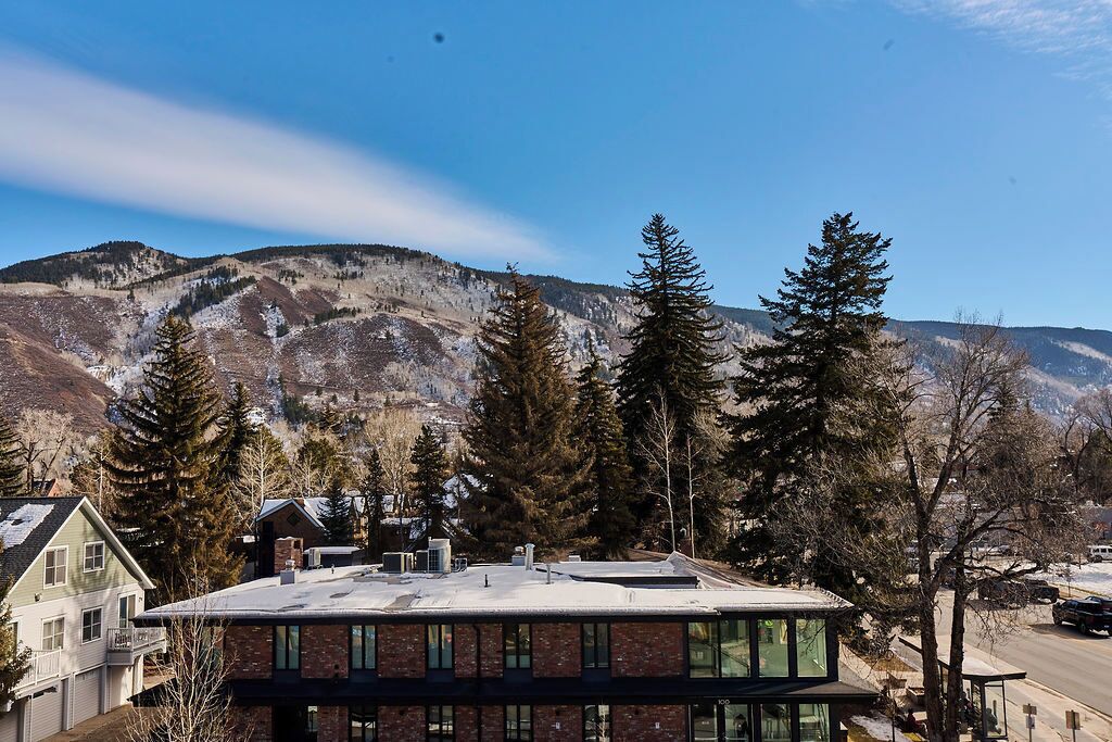 A modern building with a flat roof, surrounded by tall evergreen trees and a mountainous backdrop.