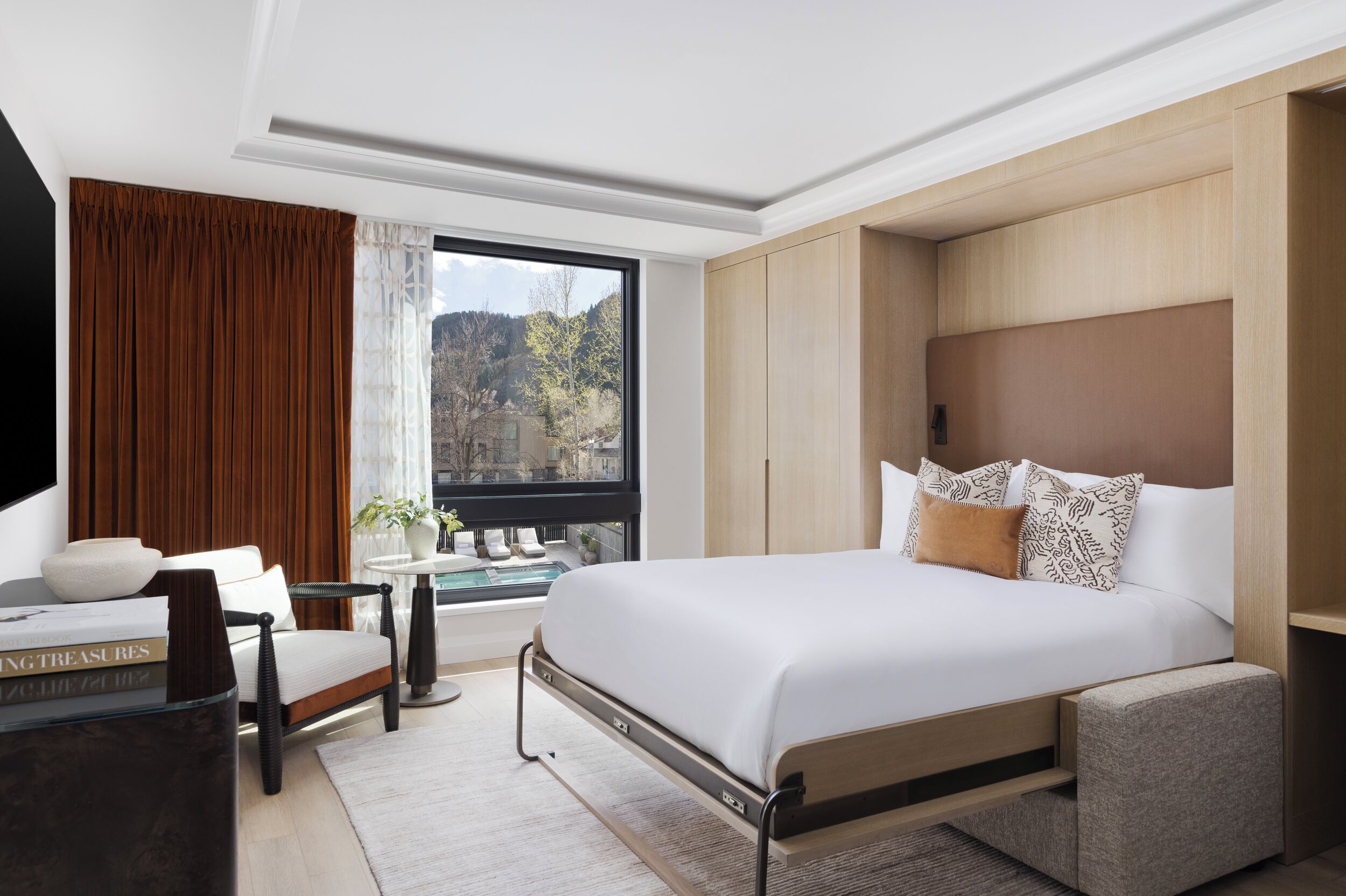 A modern hotel room with a large bed, a seating area with a chair and a small table, a view of the outdoors through a window, and built-in wooden cabinets.