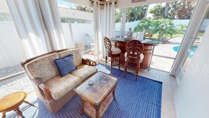 Um pátio ensolarado com um sofá de vime, uma mesa de centro de vidro e uma cadeira de madeira.