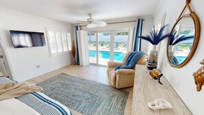 Um quarto espaçoso com uma cama grande, um espaço de estar com uma poltrona e uma mesa, uma TV fixada na parede, e vista para a piscina através das portas de correr de vidro.
