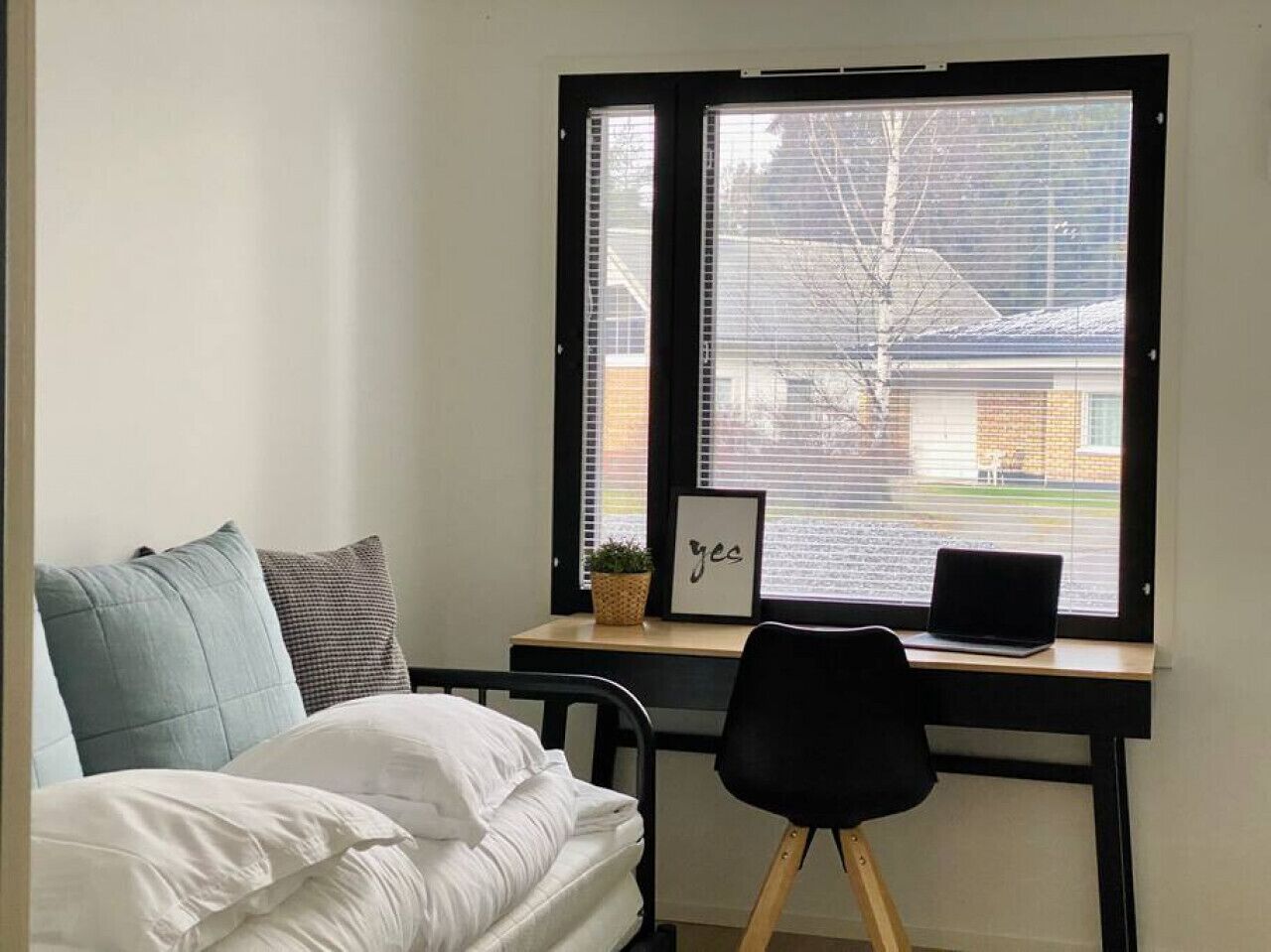 Une chambre à coucher avec un lit, un bureau avec un ordinateur portable, une chaise et une fenêtre avec des stores.