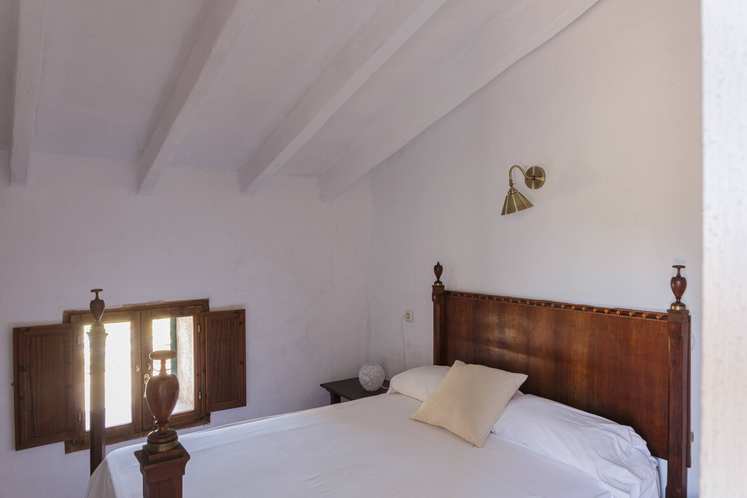 A bedroom with a wooden bed, white bedding, a nightstand, a wall-mounted lamp, and a window with shutters.