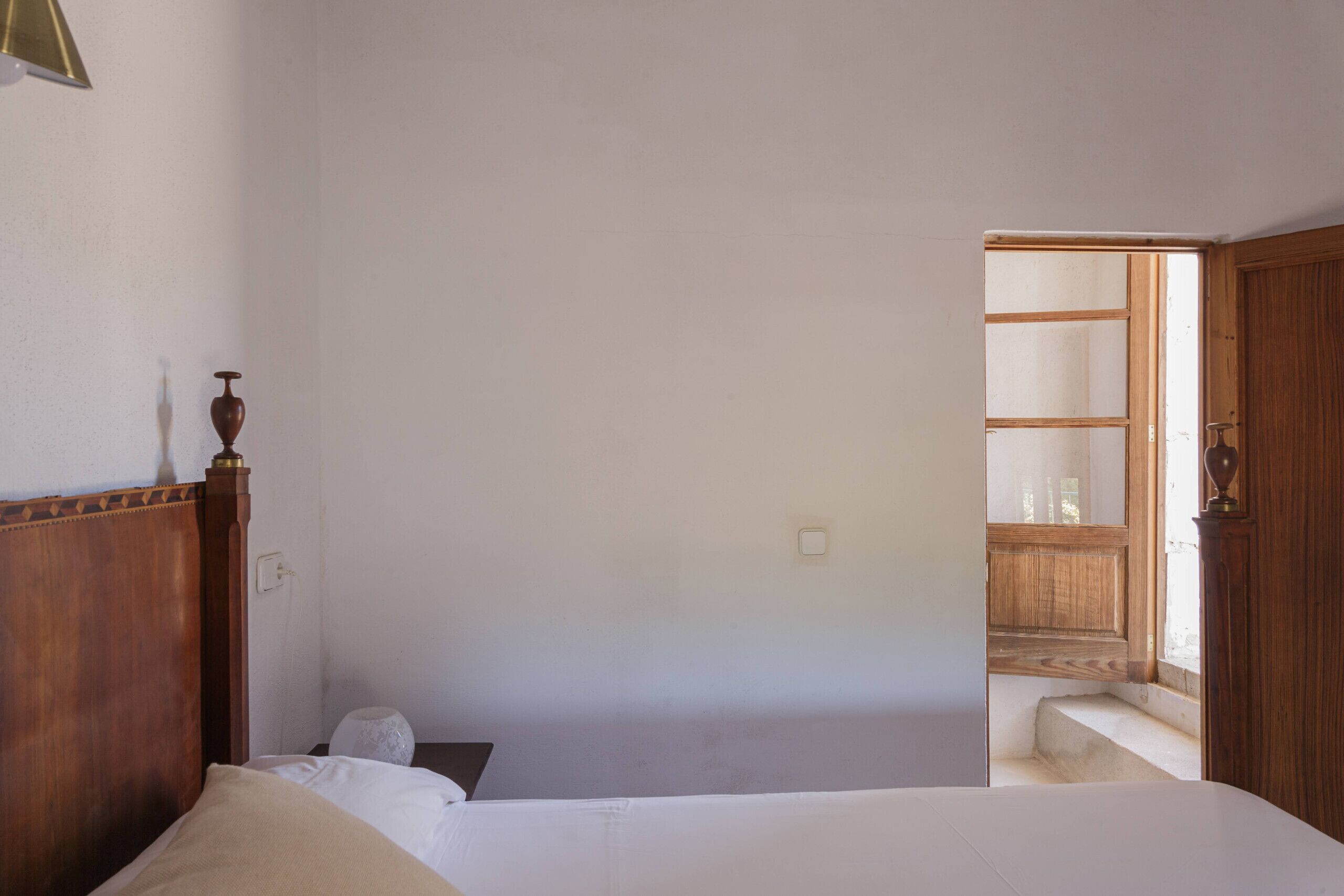 A bedroom with a wooden headboard, a bed with white linens, a bedside table, and a nightstand. There is a door leading to a hallway with a wooden door and a window.