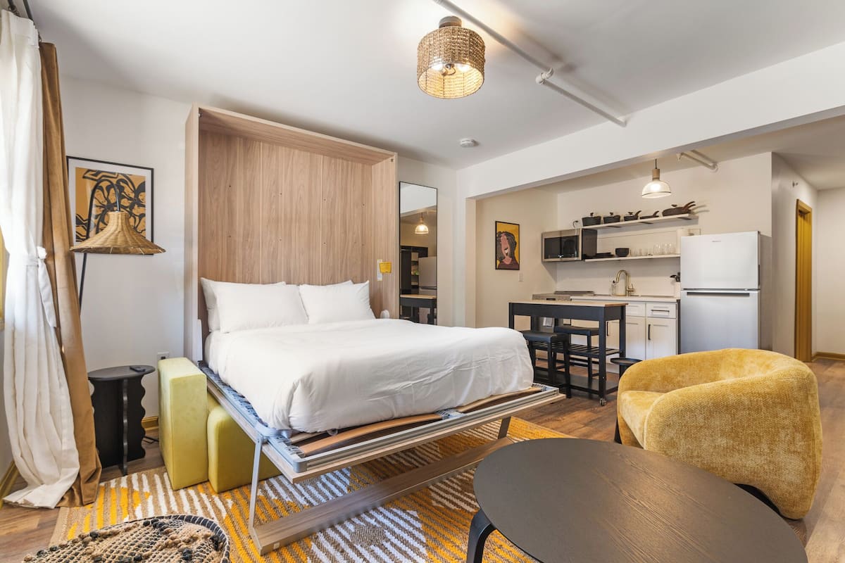 Ein kompaktes Hotelzimmer mit Bett, kleinem Schreibtisch, Stuhl, Sofa, Küchenzeile und Badezimmer.