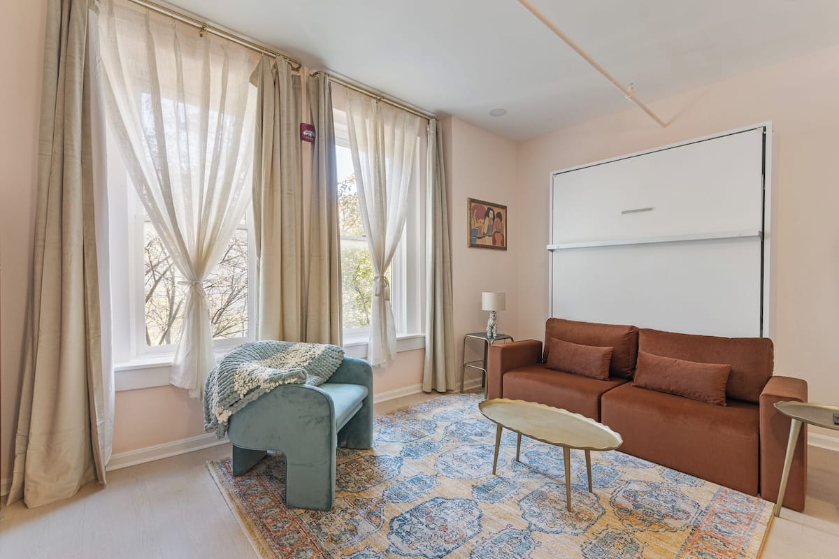 Ein Wohnzimmer mit einer Couch, einem kleinen Tisch, einem Sessel und einem großen Fenster mit Vorhängen.