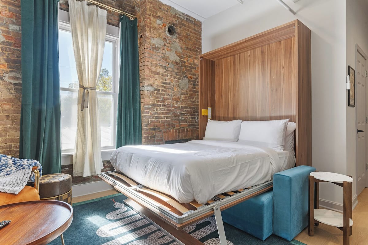 Ein modernes Schlafzimmer mit Backsteinwand, Holz-Kopfteil, Bett mit weißer Bettwäsche, einer blauen Bank, einem kleinen runden Tisch und Blick ins Freie.