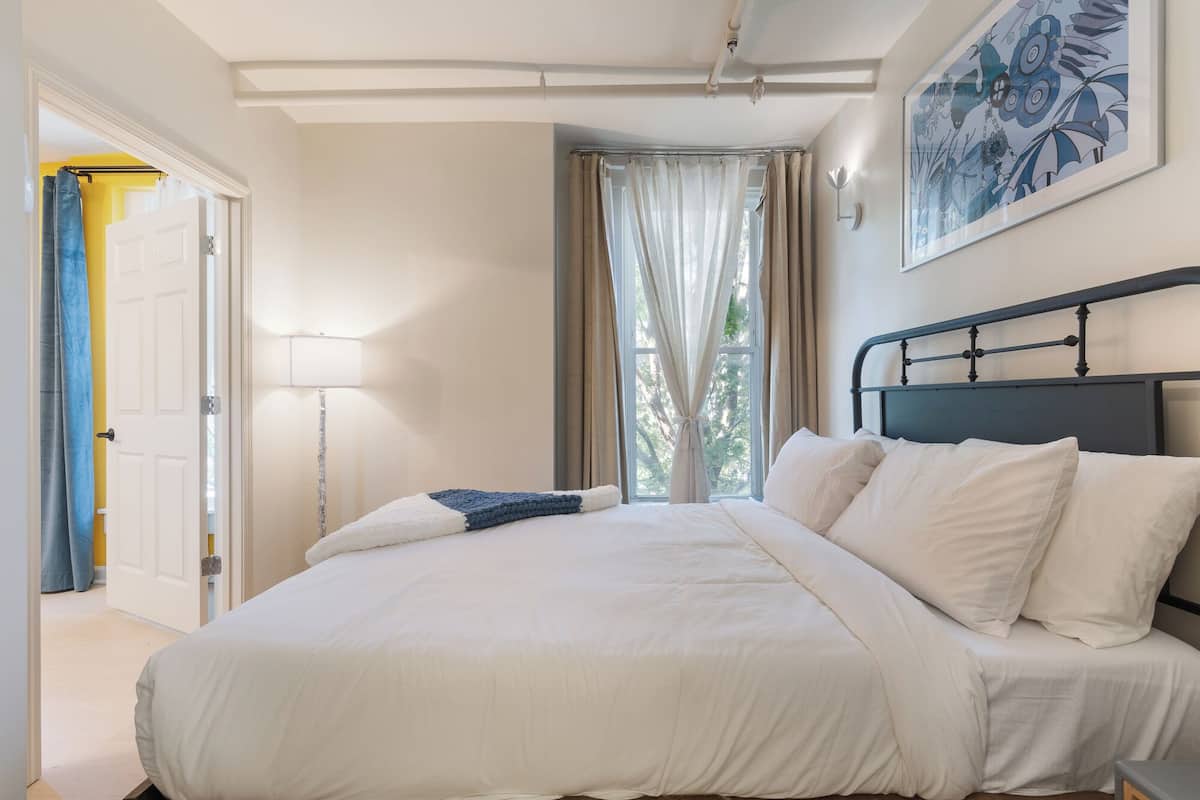 Ein ordentlich bezogenes Bett mit weißen Laken, einer Nachttischlampe und einem Fenster mit Vorhängen.