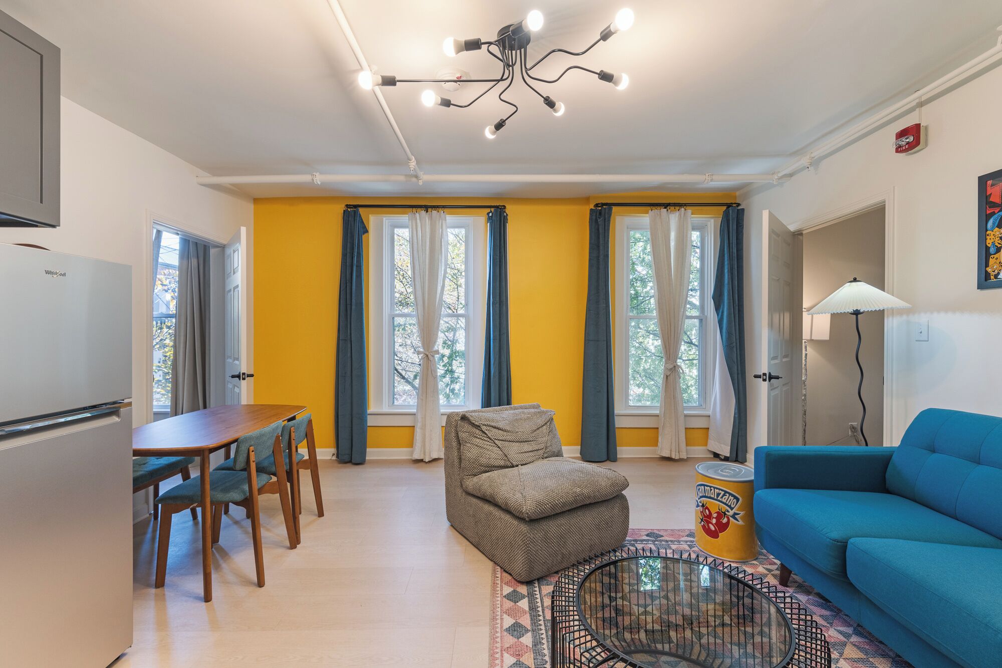 Ein Wohnzimmer mit blauem Sofa, einem Esstisch mit Stühlen, einem gemusterten Teppich und einer Deckenleuchte.