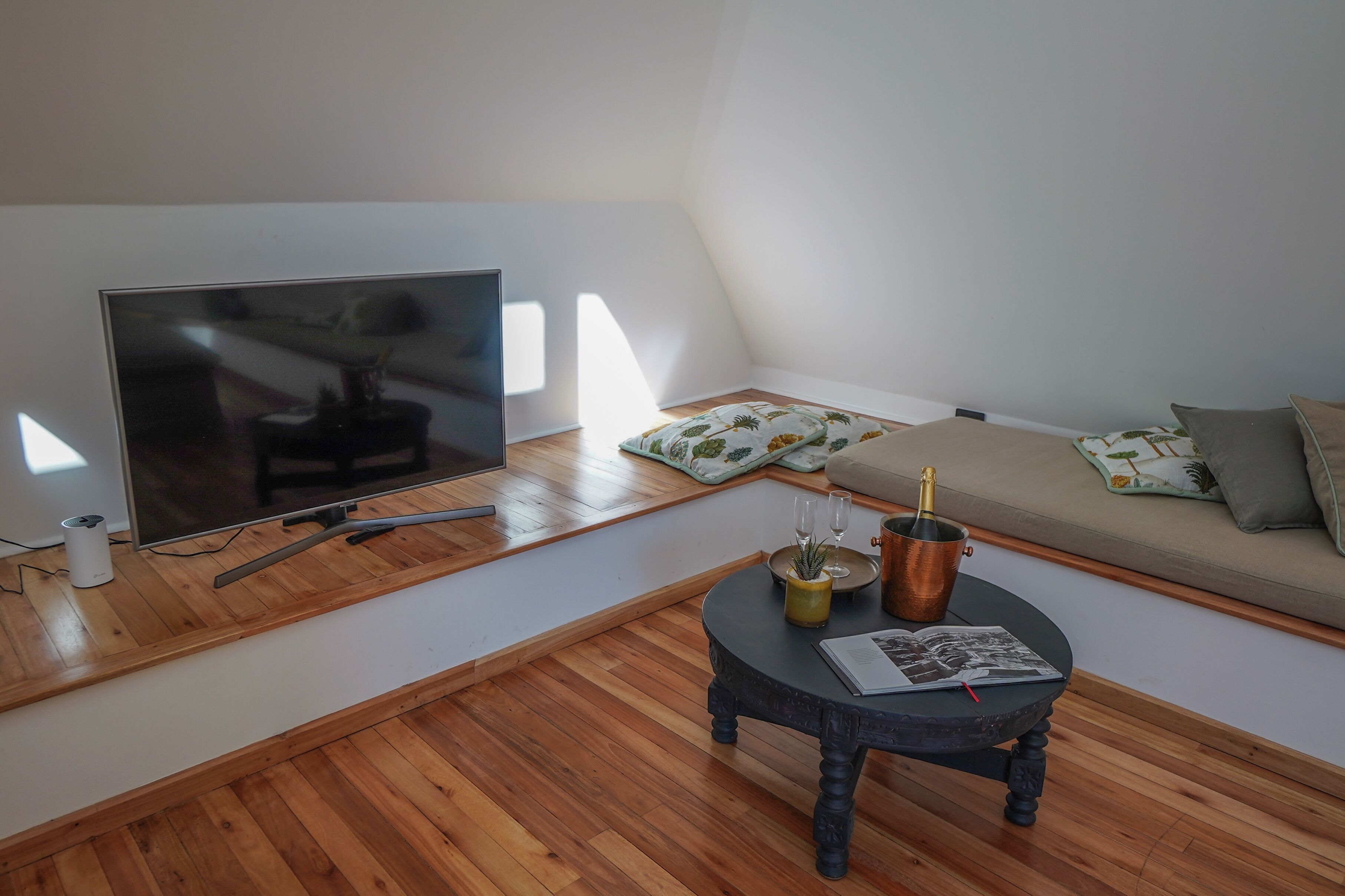A modern living room with a flat-screen TV, wooden flooring, a round coffee table with a magazine, and a sofa with patterned cushions.