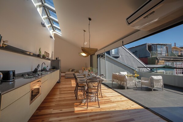 A modern kitchen with wooden flooring, a dining area with chairs, and a view of the city through large windows. - Altillo Puerto Claro (Valparaíso)