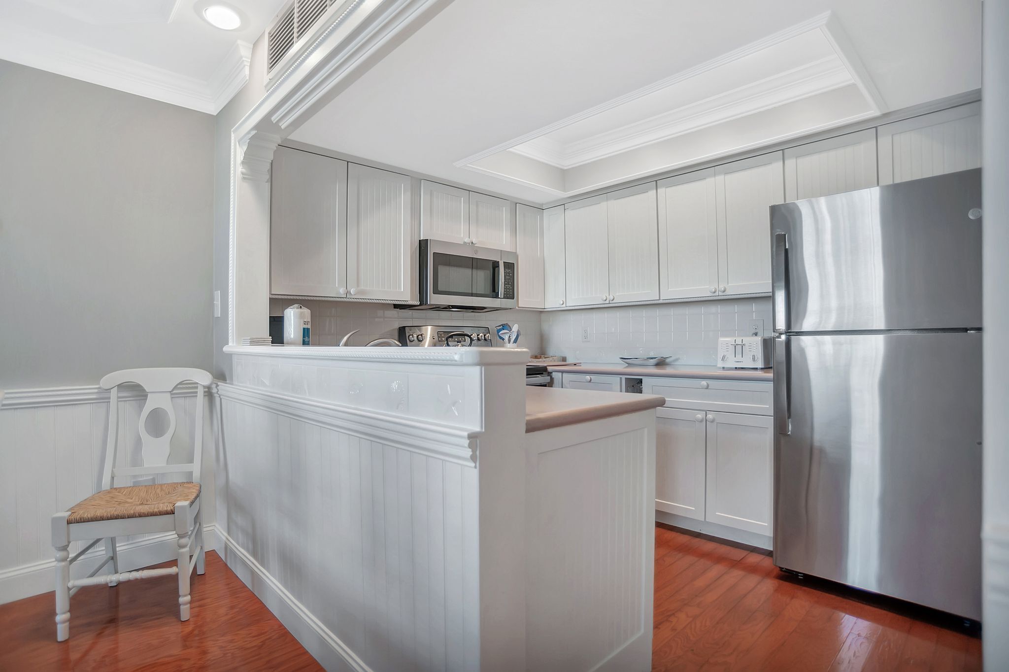 A kitchen with white cabinetry, a stainless steel refrigerator, a microwave, and a built-in island with a chair.