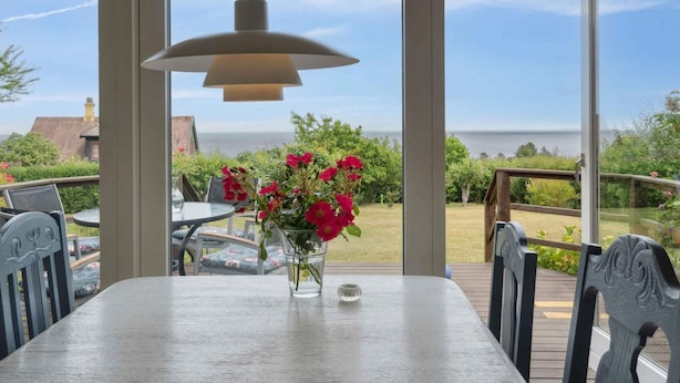 Ein Essbereich mit Tisch, Stühlen, einer Pendelleuchte und Meerblick durch große Fenster.