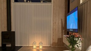 A cozy room with a TV mounted on the wall, a small table with a vase of flowers, and two lit lanterns on the floor. - 5 Star Holiday Home in Eskebjerg (Eskebjerg)