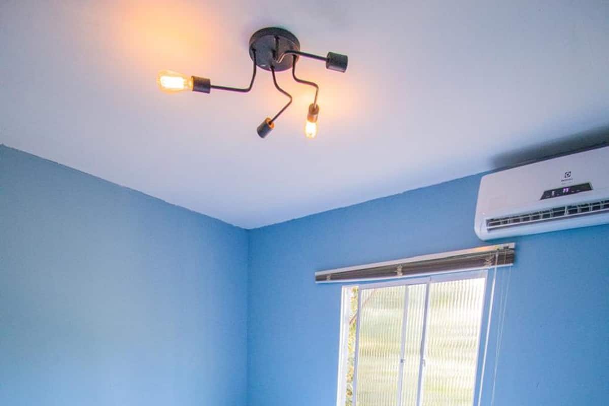 Un dormitorio con cama, mesita de noche, ventana con persianas, unidad de aire acondicionado e iluminación en el techo.