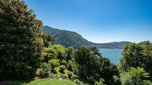 Um jardim exuberante com diversas árvores e arbustos, céu azul límpido e vista distante de um lago e montanhas.