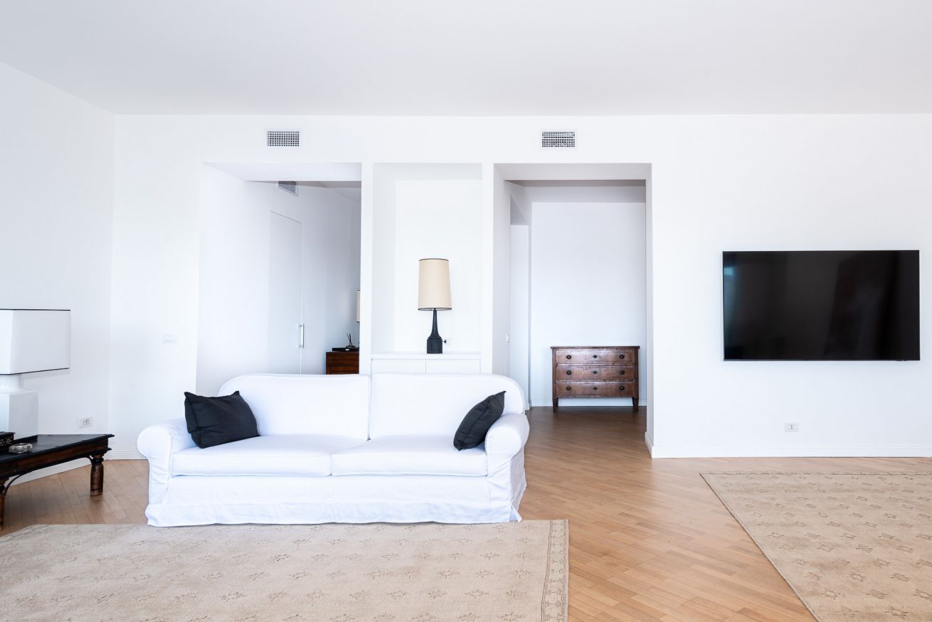 Un soggiorno minimalista con un divano bianco, un tavolino in legno, una lampada, una TV e un tappeto decorato.