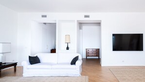 Uma sala de estar minimalista com um sofá branco, uma mesa de centro de madeira, uma luminária, uma televisão e um tapete estampado.