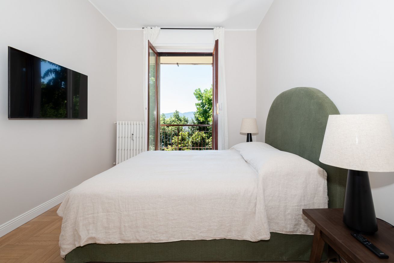 Una camera da letto con un letto, un comodino, una lampada, una TV appesa al muro e vista sul verde attraverso una porta aperta.
