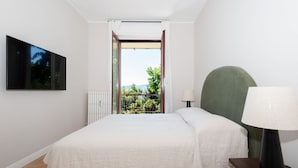 Quarto com cama, mesa de cabeceira, luminária, TV na parede e vista para o verde através de uma porta aberta.