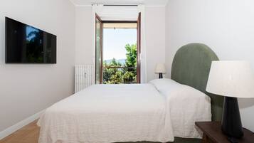 Una camera da letto con un letto, un comodino, una lampada, una TV appesa al muro e vista sul verde attraverso una porta aperta.